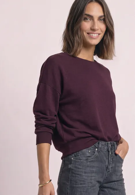 Koszulka z długim rękawem i elastycznym wykończeniem rustic rouge mel. Uśmiechnięta kobieta w bordowym swetrze i szarych dżinsach na jasnym tle.