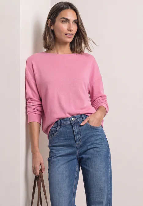 Koszulka z długim rękawem i elastycznym wykończeniem warm blush mel. Kobieta w różowym swetrze i niebieskich dżinsach trzyma torbę.
