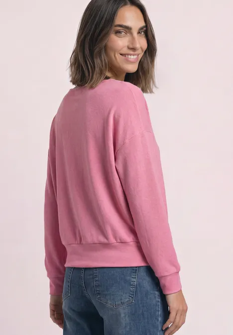 Koszulka z długim rękawem i elastycznym wykończeniem warm blush mel. Kobieta z włosami do ramion ma różowy sweter i niebieskie dżinsy, uśmiecha się patrząc przez ramię.