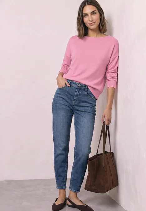Koszulka z długim rękawem i elastycznym wykończeniem warm blush mel. Kobieta w różowym swetrze, niebieskich dżinsach i czarnych balerinach z brązową torbą.