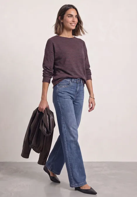 Une femme souriante porte un haut rayé à manches longues, un jean bleu et tient une veste.