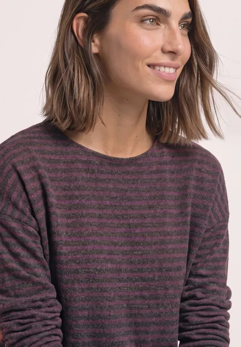 Une femme aux cheveux mi-longs porte un pull à rayures violet et noir à manches longues, sourire discret.