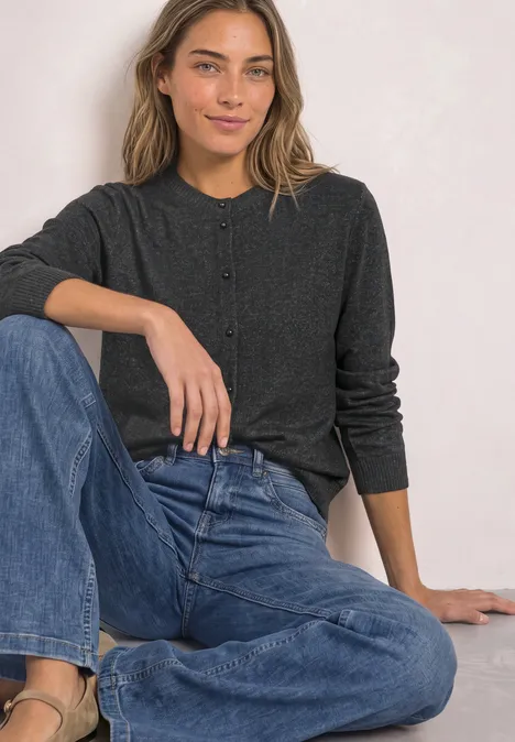 Eine lächelnde Frau in dunkelgrauem Cardigan und blauen Jeans sitzt entspannt vor hellem Hintergrund.