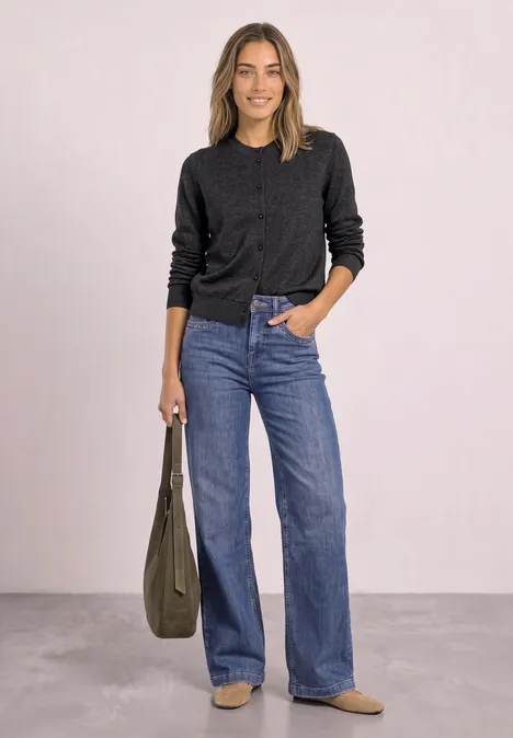 Eine Frau trägt schwarzen Pullover, weite Jeans, Boots und hält eine grüne Tasche.