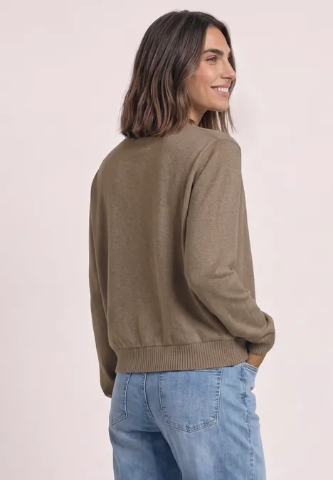 Une femme en jean bleu et pull olive regarde par-dessus son épaule en souriant.