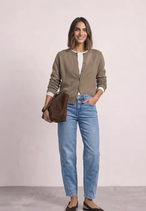 Femme souriante en cardigan beige, top blanc et jean bleu, tenant une pochette marron sur fond clair.