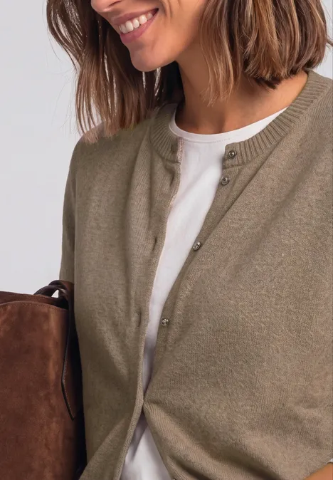 Femme aux cheveux bruns mi-longs porte un cardigan kaki sur un t-shirt blanc et tient un sac marron.