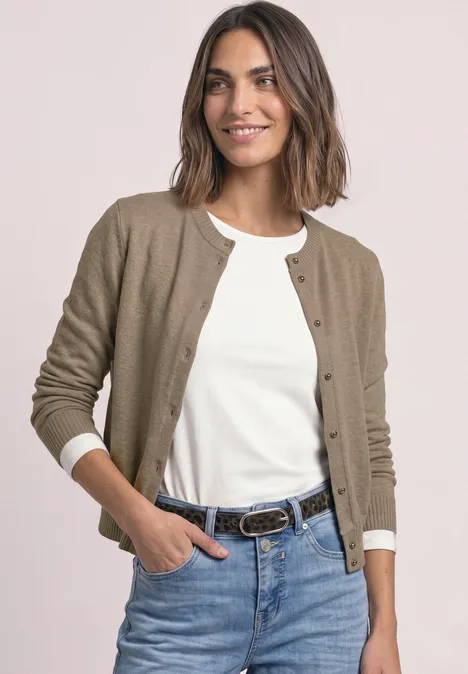 Une femme souriante en jean, t-shirt blanc et gilet marron devant un fond clair.