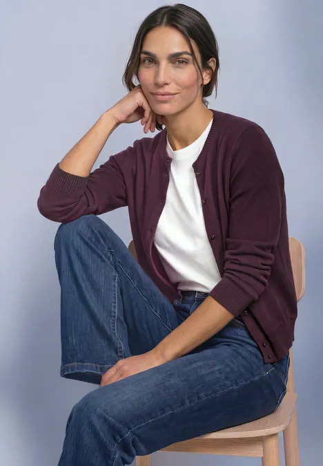 Frau im lila Cardigan, weißem T-Shirt und Jeans sitzt lässig auf einem Hocker und stützt den Kopf auf die Hand.