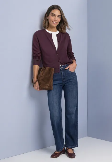Frau lächelt, trägt weiße Bluse, violette Jacke, gerade Jeans, braune Loafers und hält eine braune Clutch.