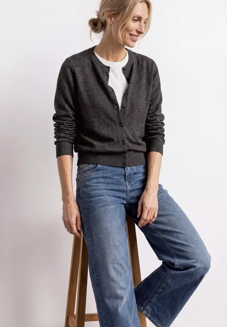 Femme souriante appuyée sur un tabouret, en cardigan gris, t-shirt blanc et jean large bleu.