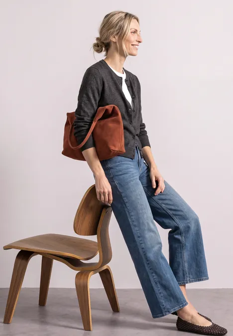 Femme en cardigan gris, chemise blanche et jean large, portant un sac en cuir marron et posant près d’une chaise en bois.