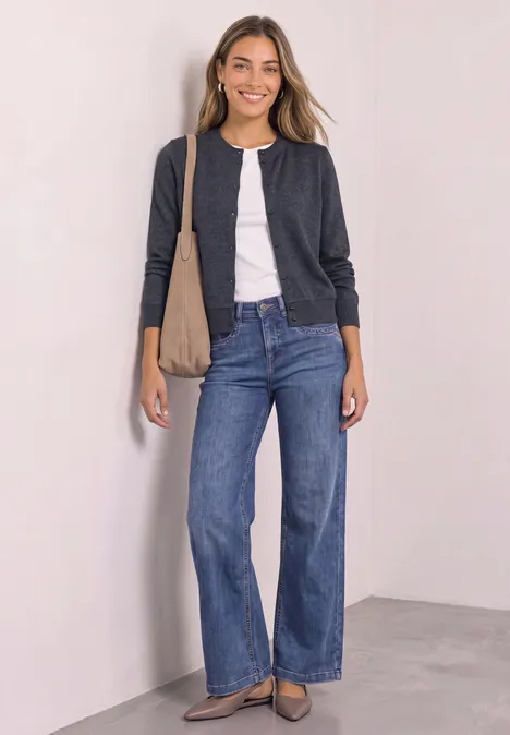 Frau lächelt in Jeans, weißem Shirt und dunkelgrauer Jacke mit Schultertasche gegen weiße Wand.