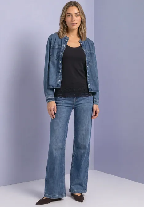 Femme en chemisier noir, veste en jean et jean évasé devant un fond neutre.