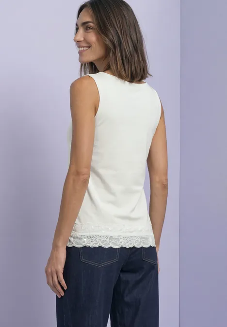 Femme en top blanc sans manches à bordure en dentelle et jean foncé souriant devant un mur violet.
