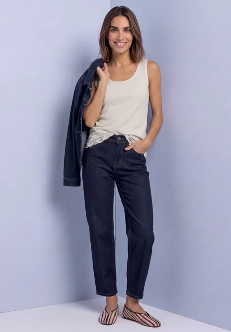 Femme en top crème et jean sombre, souriante, tenant une veste sur l’épaule.