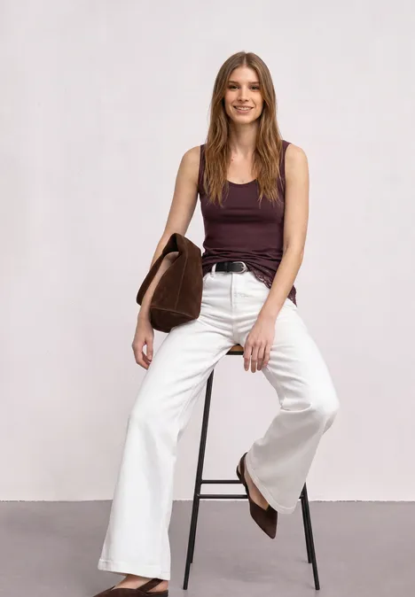 Top mit Rundhals und Spitzenkante rustic rouge Frau mit braunem Top und weißer Schlaghose sitzt lächelnd auf einem Hocker und hält eine braune Tasche.