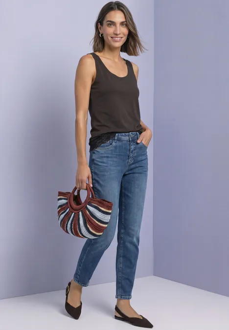 Femme en débardeur noir et jean, tenant un sac coloré en forme de demi-lune, posant devant un mur gris.