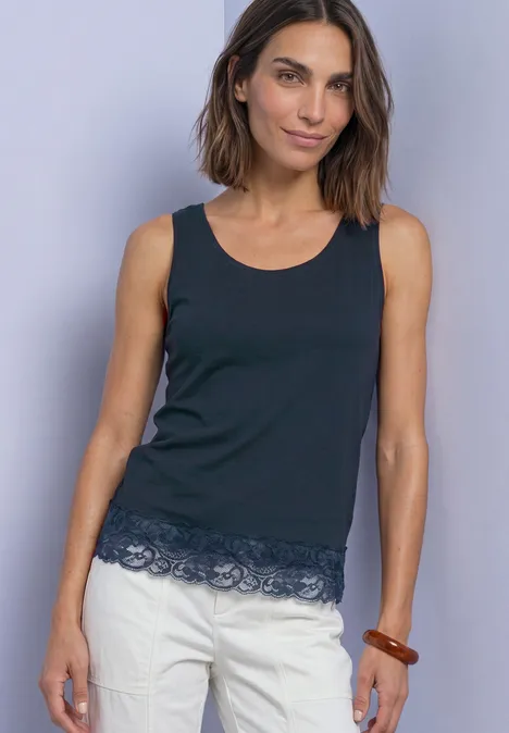 Femme portant un débardeur noir sans manches à bordure en dentelle et un pantalon blanc devant un mur gris clair.