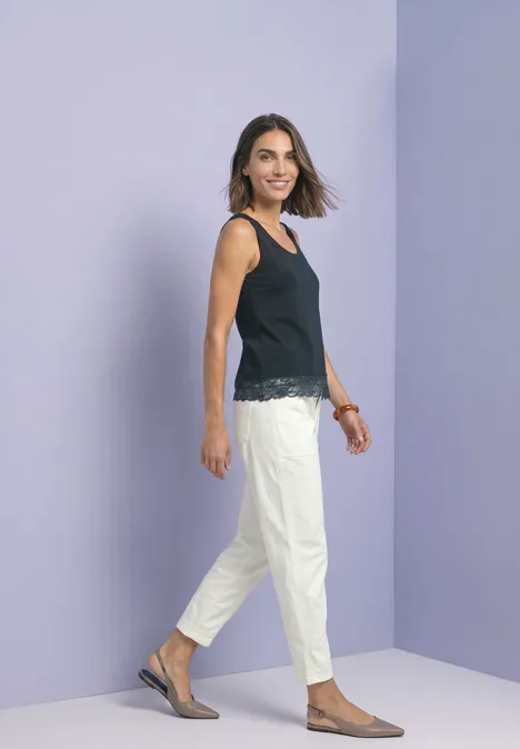 Femme souriante en top noir et pantalon blanc devant un mur violet.