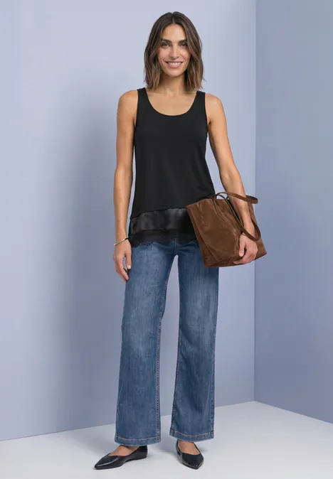 Top met ronde hals en decoratieve kant Black Vrouw in zwart mouwloos topje met volant, wijde jeans en bruine tas, glimlachend voor een grijze achtergrond.