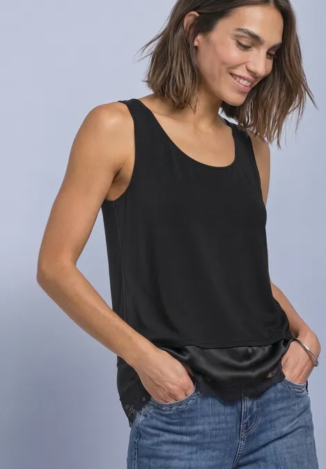 Top met ronde hals en decoratieve kant Black Vrouw in zwarte mouwloze top met satijnen zoom en blauwe jeans, glimlachend met handen in de zakken.