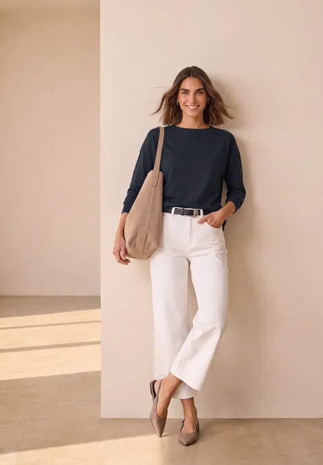 3/4-Arm Shirt mit Rippstrukturdetails indigo night Eine Frau lehnt an einer beigen Wand, trägt schwarzen Pullover, weiße Culottes, beige Tasche und Schuhe, lächelt.