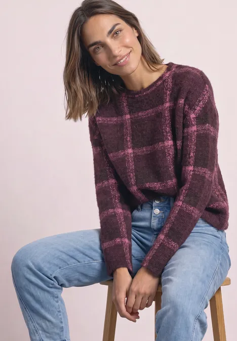 Frau auf Hocker sitzt, trägt karrierten bordeauxroten Pullover und blaue Jeans, lächelt leicht.