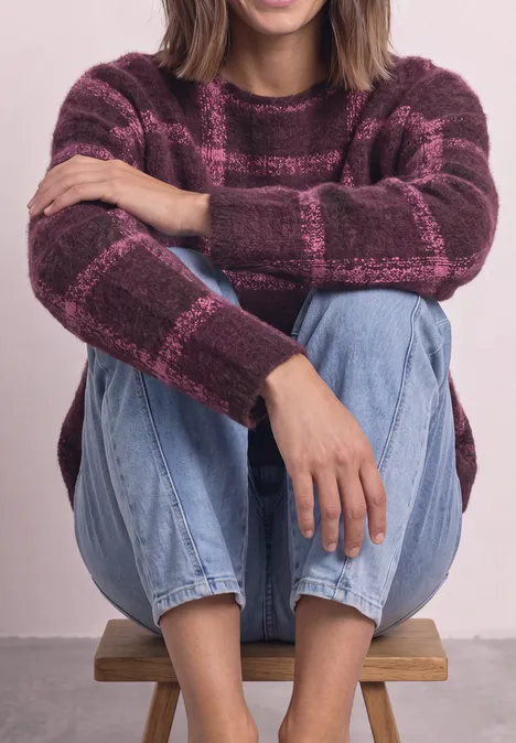 Frau sitzt auf einem Holzhocker, trägt einen lila-rosa karierten Pullover und blaue Jeans, Arme um die Knie geschlungen.