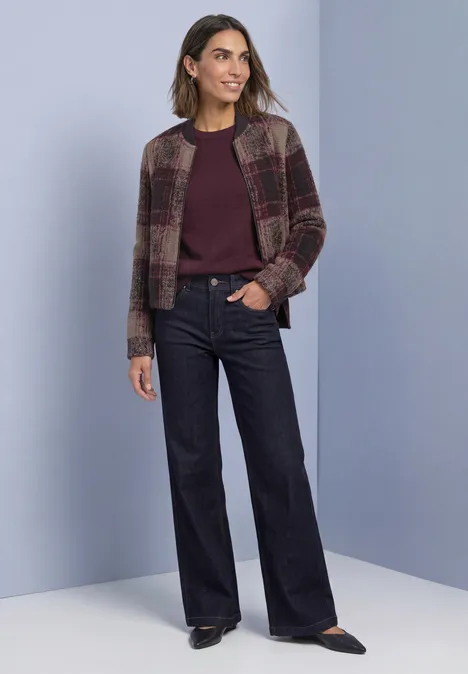 Frau steht vor hellem Hintergrund, trägt lila Pullover, karierte Jacke und weite dunkle Jeans.