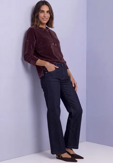 Femme souriante appuyée contre un mur mauve, porte un haut bordeaux, un jean foncé et escarpins noirs.