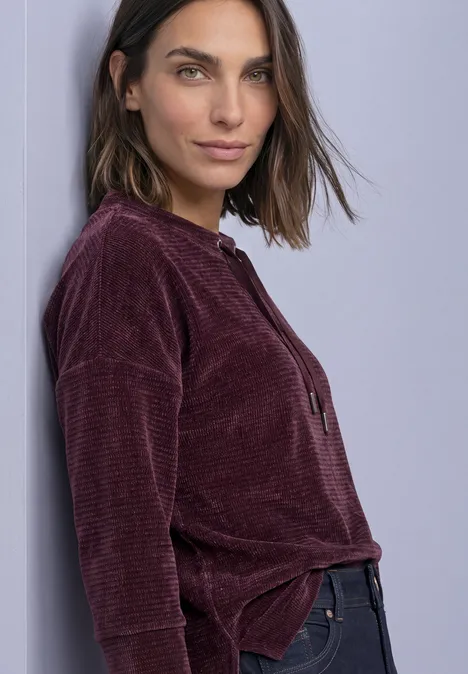 Femme appuyée contre un mur, pull en velours côtelé violet et jean, regardant l'appareil photo.