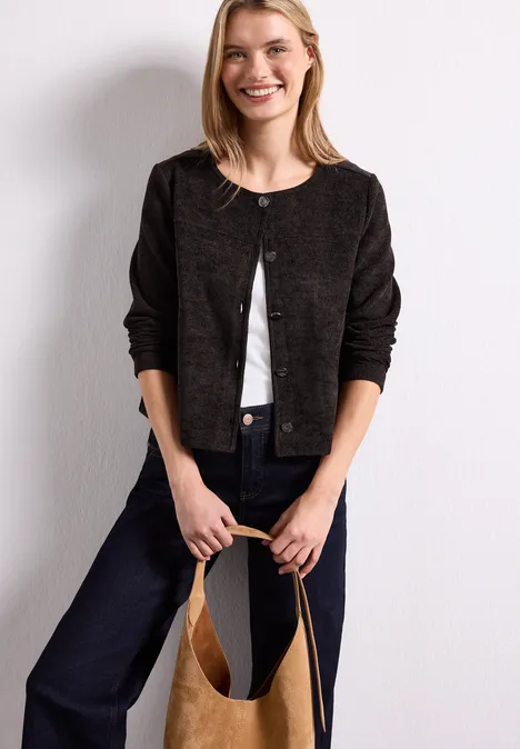 Veste à manches longues, col rond et boutons black coffee Frau lehnt an weißer Wand, trägt schwarzen Cardigan, weiße Bluse, Jeans und hält eine beige Tasche.