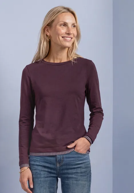 Femme blonde souriante en tee-shirt à manches longues violet et jean, main dans la poche, regard de côté.