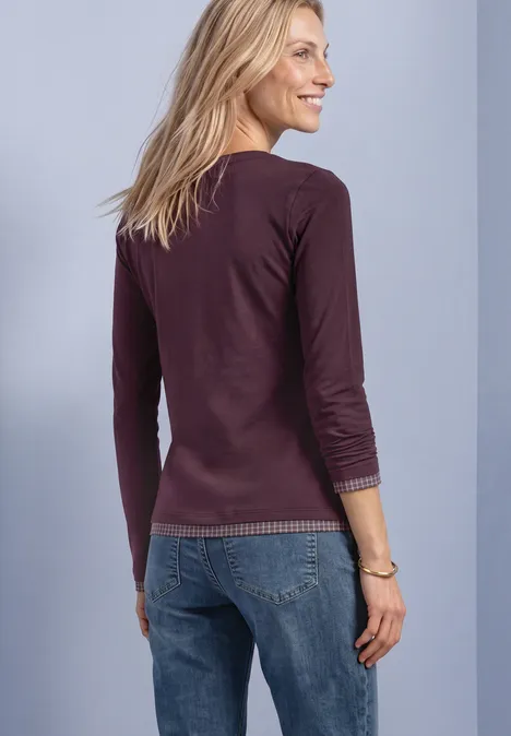 Une femme blonde vue de dos, portant un haut violet à manches longues et un jean bleu, souriant.