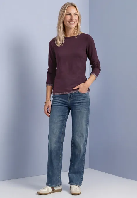 Une femme en jean, pull bordeaux et baskets blanches devant un fond bleu clair.
