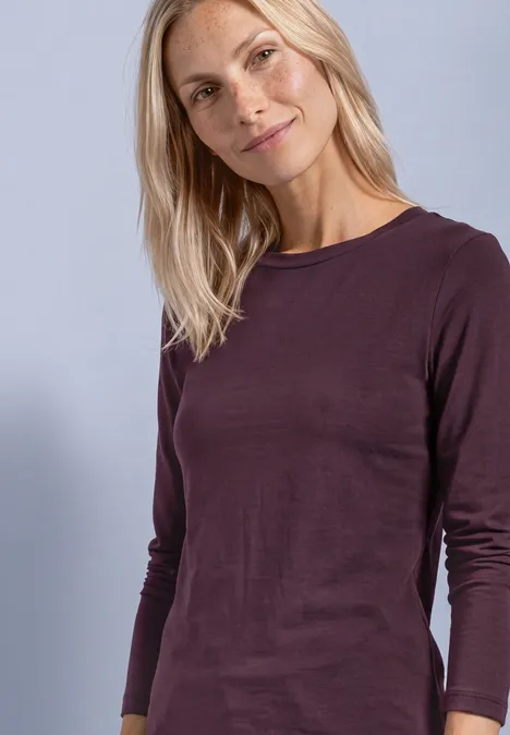 Une femme blonde souriante porte un t-shirt à manches longues prune sur fond parme.