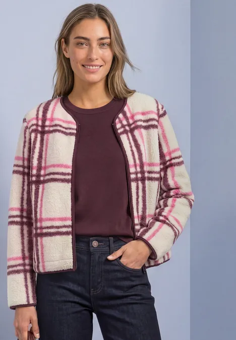 Frau trägt weiß-rosa-karierte Jacke, weinrotes Shirt und dunkle Jeans vor hellem Hintergrund.