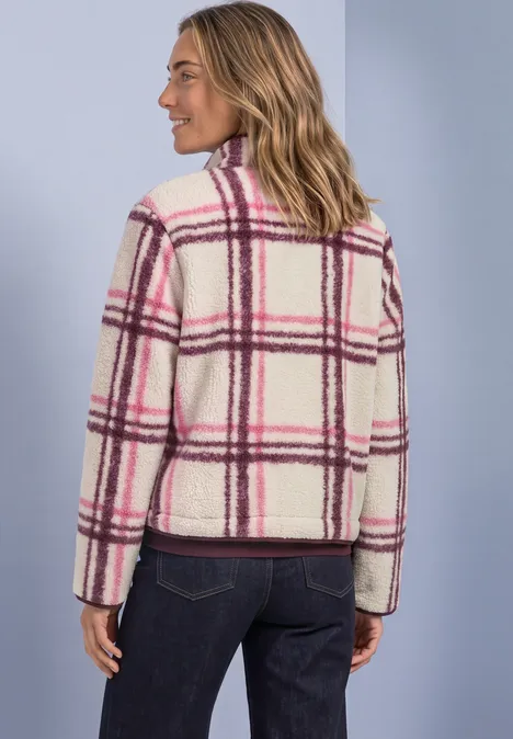 Frau trägt weiße, flauschige Jacke mit pink-lila Karomuster, steht seitlich vor hellblauem Hintergrund.