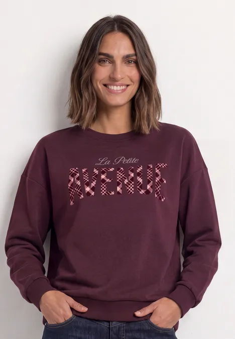 Koszulka z długim rękawem z nadrukiem graficznym rustic rouge Kobieta ma bordową bluzę z kwiatowym haftem i napisem.
