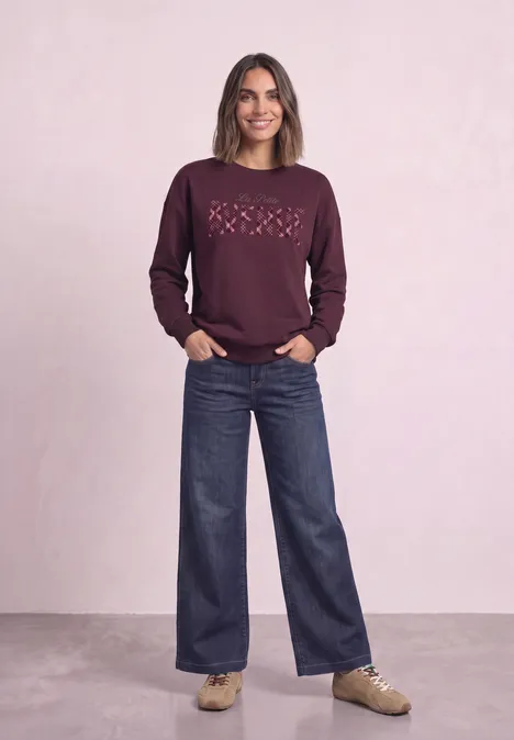 Koszulka z długim rękawem z nadrukiem graficznym rustic rouge Kobieta w bordowej bluzie z serduszkami, szerokich dżinsach i beżowych trampkach na jasnym tle.