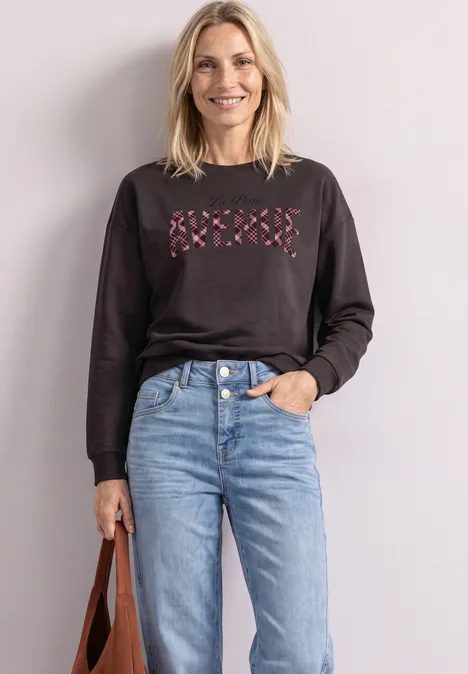 Femme souriante en sweat noir à motif croix roses, jean bleu clair, tenant un sac à main marron.