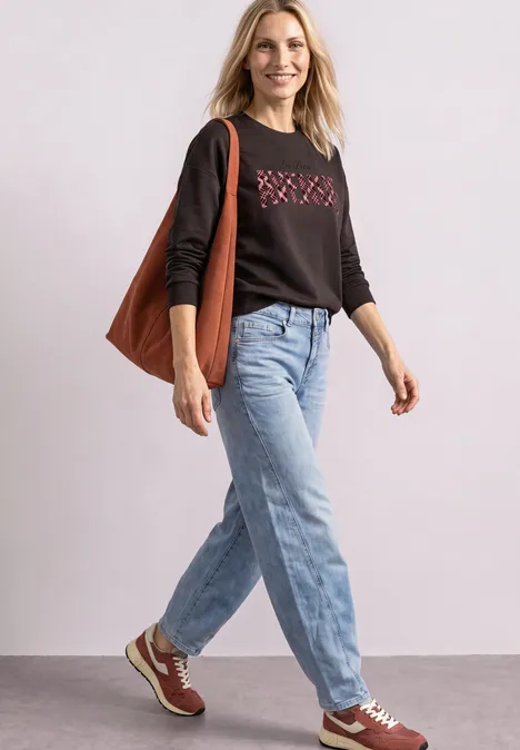 Femme en jean, pull noir et baskets, portant un sac marron, marchant en souriant.
