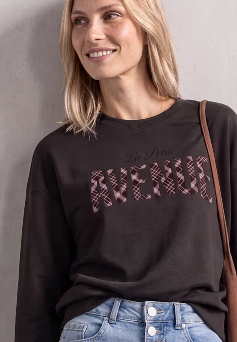 Une femme souriante porte un pull noir avec lettrage zébré "AVENUE" et un jean bleu.