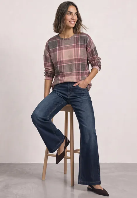 Frau mit kariertem Pullover und ausgestellten Jeans lehnt lässig an einem Hocker.