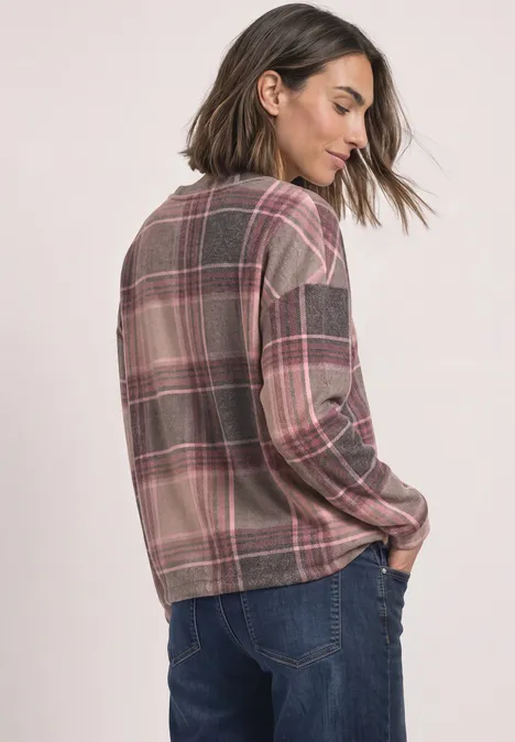 Frau in kariertem Langarmshirt in Braun- und Rosatönen, Rückenansicht, Hände in Jeanshosentaschen gesteckt.