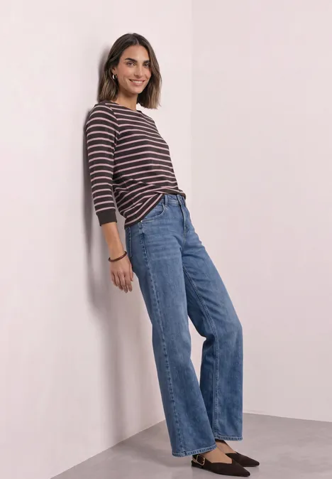 Koszulka z rękawami 3/4, dekoltem w łódkę i w paski vintage blush Uśmiechnięta kobieta w paskowanej bluzce i jeansach opiera się o ścianę.