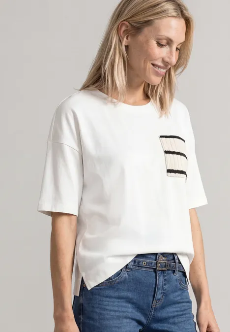 Frau in weißem T-Shirt mit gestreiftem Brusttaschen-Detail und blauen Jeans, lächelnd seitlich stehend