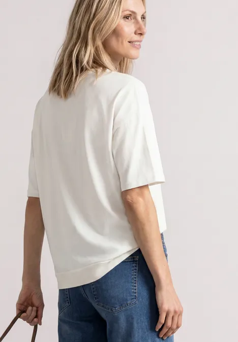Frau in weißem T-Shirt und Jeans steht seitlich und hält eine Tasche in der Hand.
