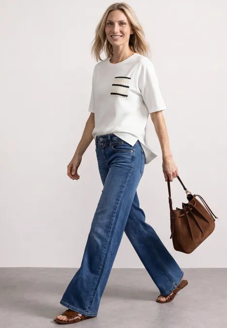 Frau in weißem T-Shirt mit schwarzem Streifendetail, weiten Jeans und brauner Tasche, die lächelnd geht.